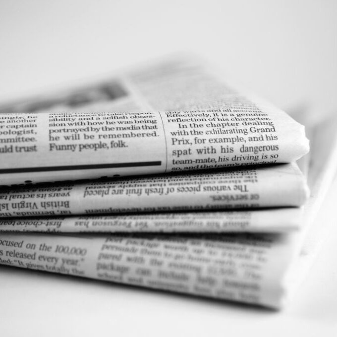 A close up black and white image of a stack four broadsheet newspapers with English text, casually piled up on a plain white background. The macro image is shot with a shallow depth of field, with only generic text visible. With copyspace.